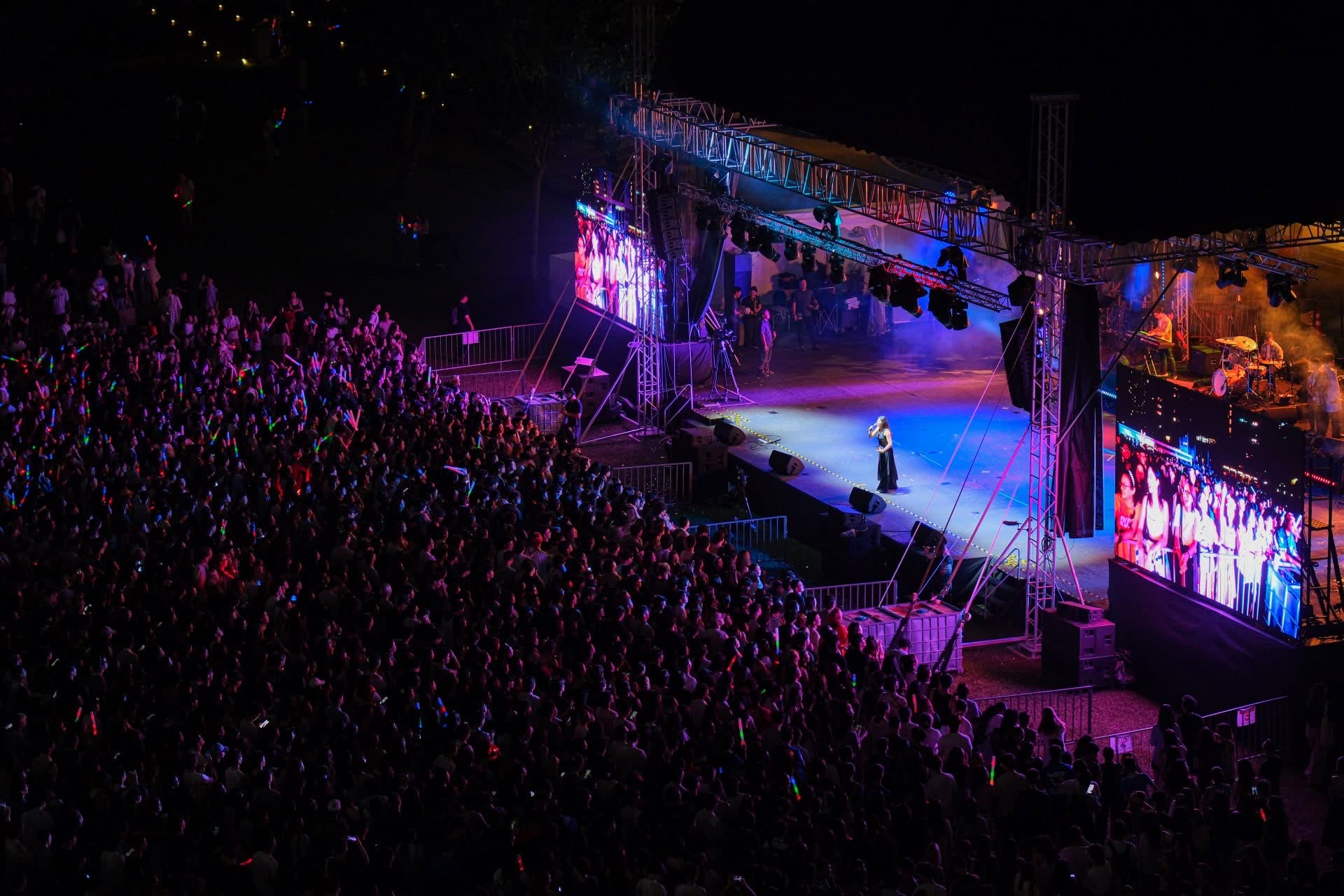A crowd at an outdoor night concert, facing a lit stage and screens